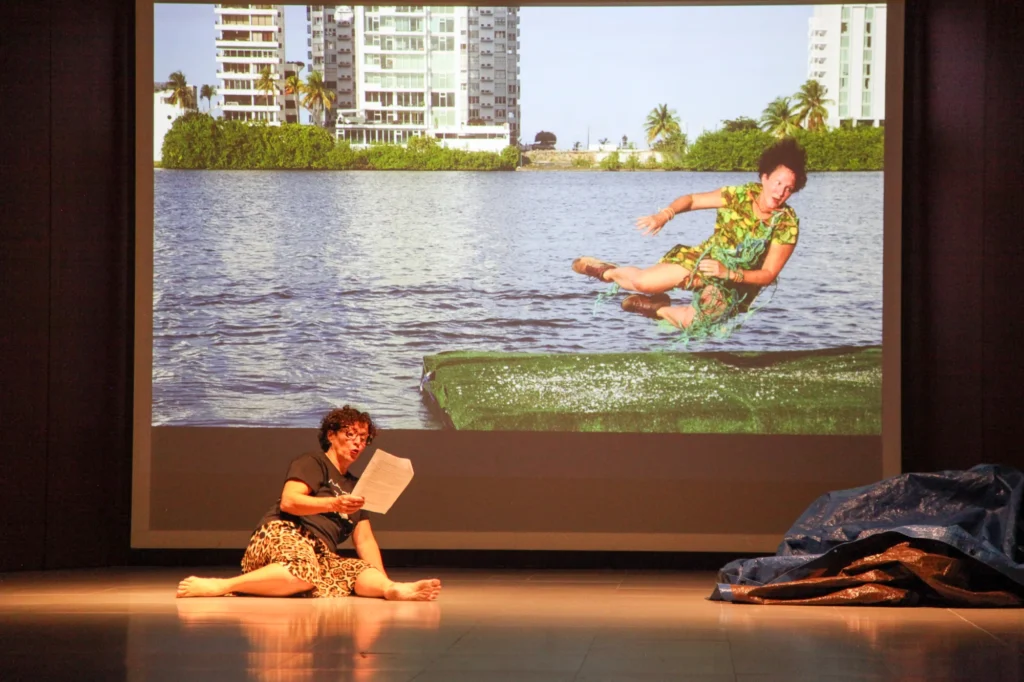 A person sits on a stage reading from a paper in front of a large screen showing an image of a woman jumping over water near blocks of flats.