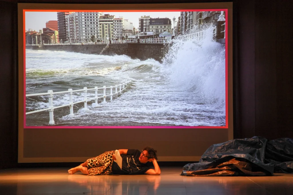 A person lies on a stage floor reading, in front of a large screen showing waves crashing against a city sea wall.
