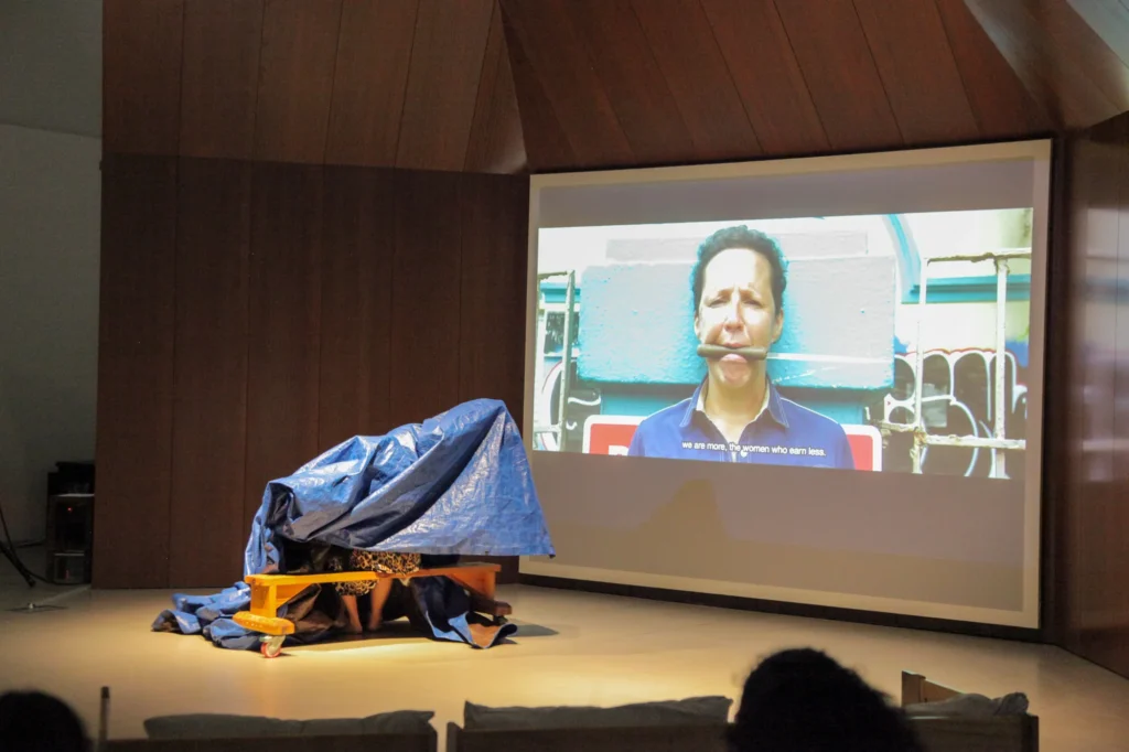 A person sits under a blue tarpaulin on a stage in front of a projection screen showing a woman with a knife in her mouth and the subtitle, “we are more, the women who earn less.”