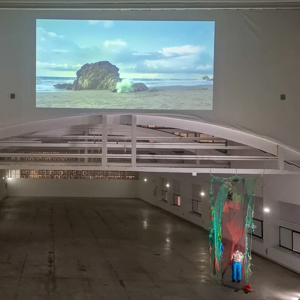 A large gallery space features a projected video of a beach on the wall and a person standing next to a hanging sculpture made of nets and translucent fabric.