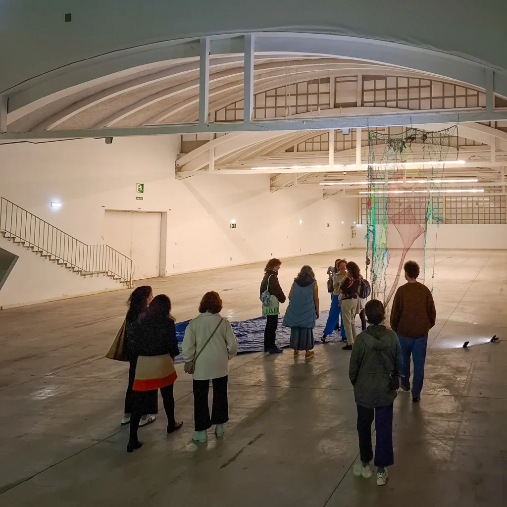 A group of people standing in a large, empty industrial hall, looking at a hanging art installation of colourful netting.