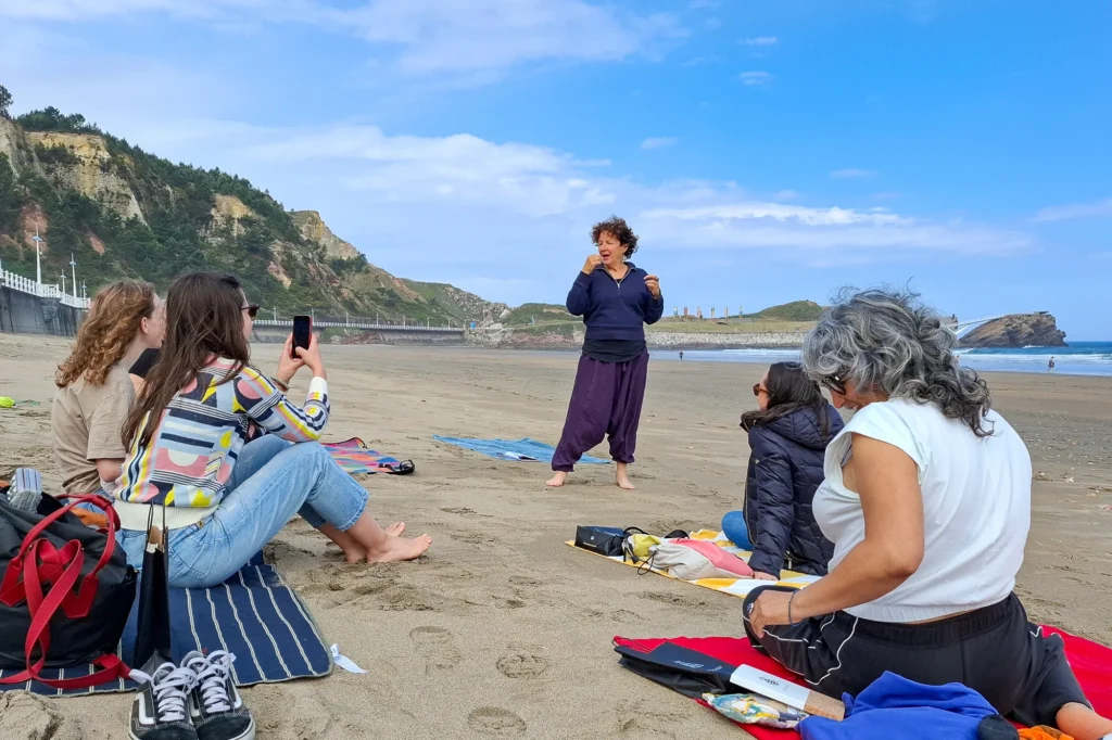 A woman talks to a group on a beach as one person photographs her with a mobile.