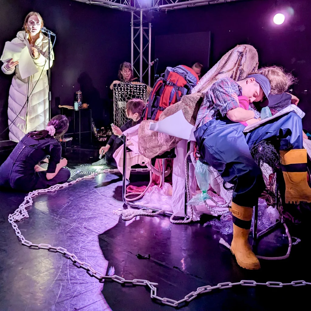 A performer reads from a piece of paper while others sit amongst props including a plastic chain, rucksacks, and yellow wellington boots on a stage.