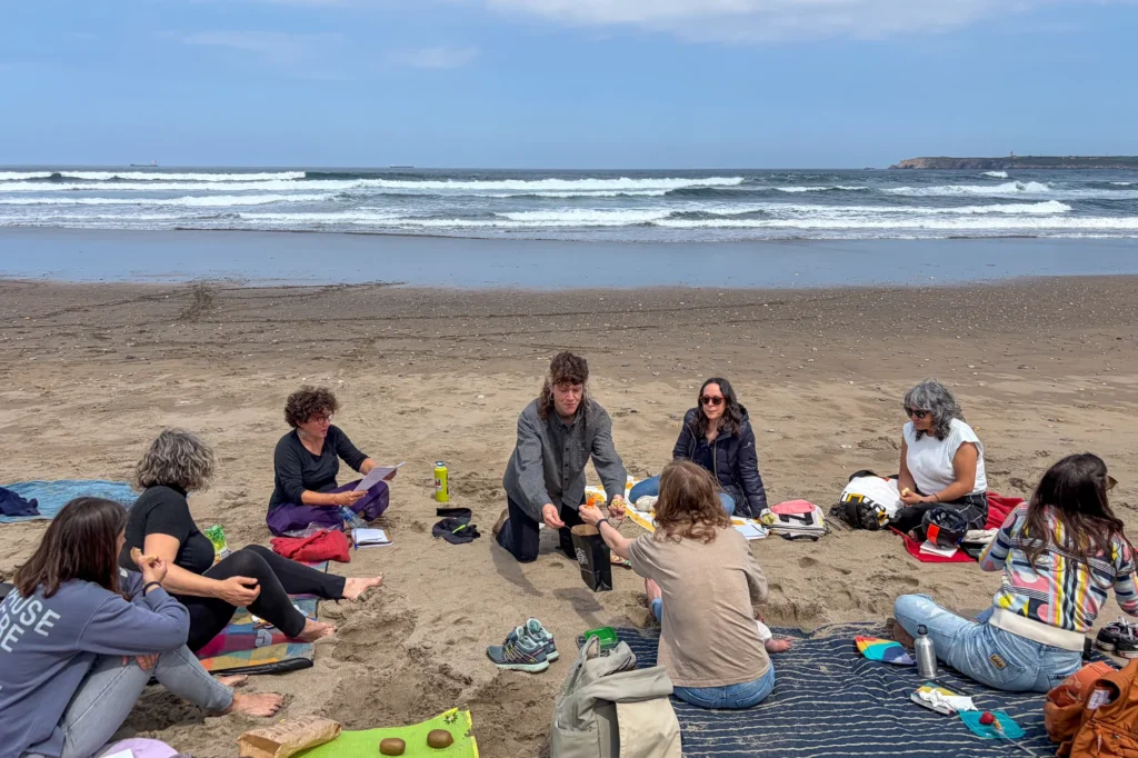 A group of people sitting in a circle on a sandy beach by the sea.