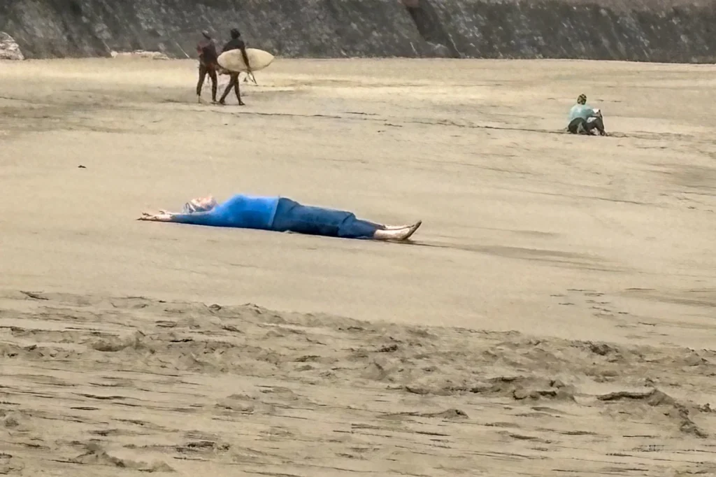 A person in a blue shirt and jeans lies flat on their back on a sandy beach