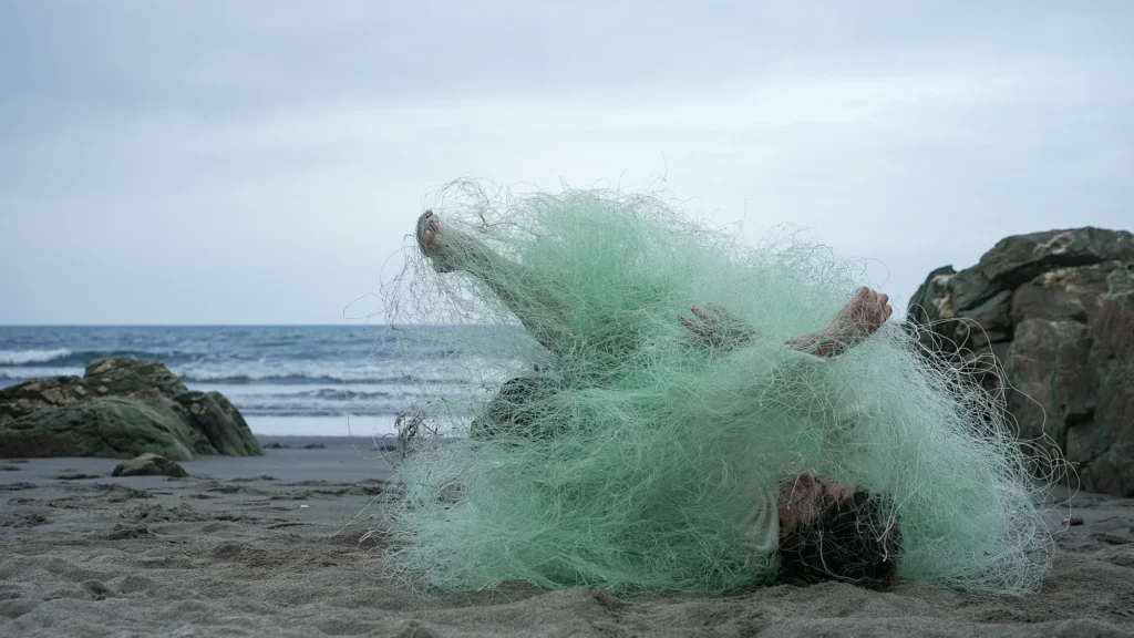 A person lying on a sandy beach, tangled in a large green fishing net.