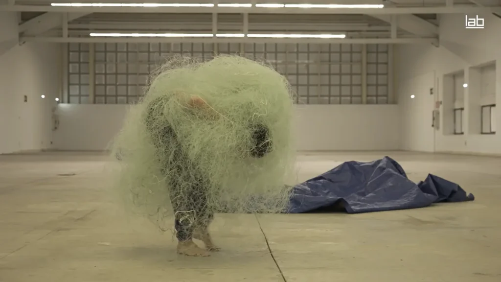 A person bent over, covered in a large mass of green netting, in an empty warehouse space.
