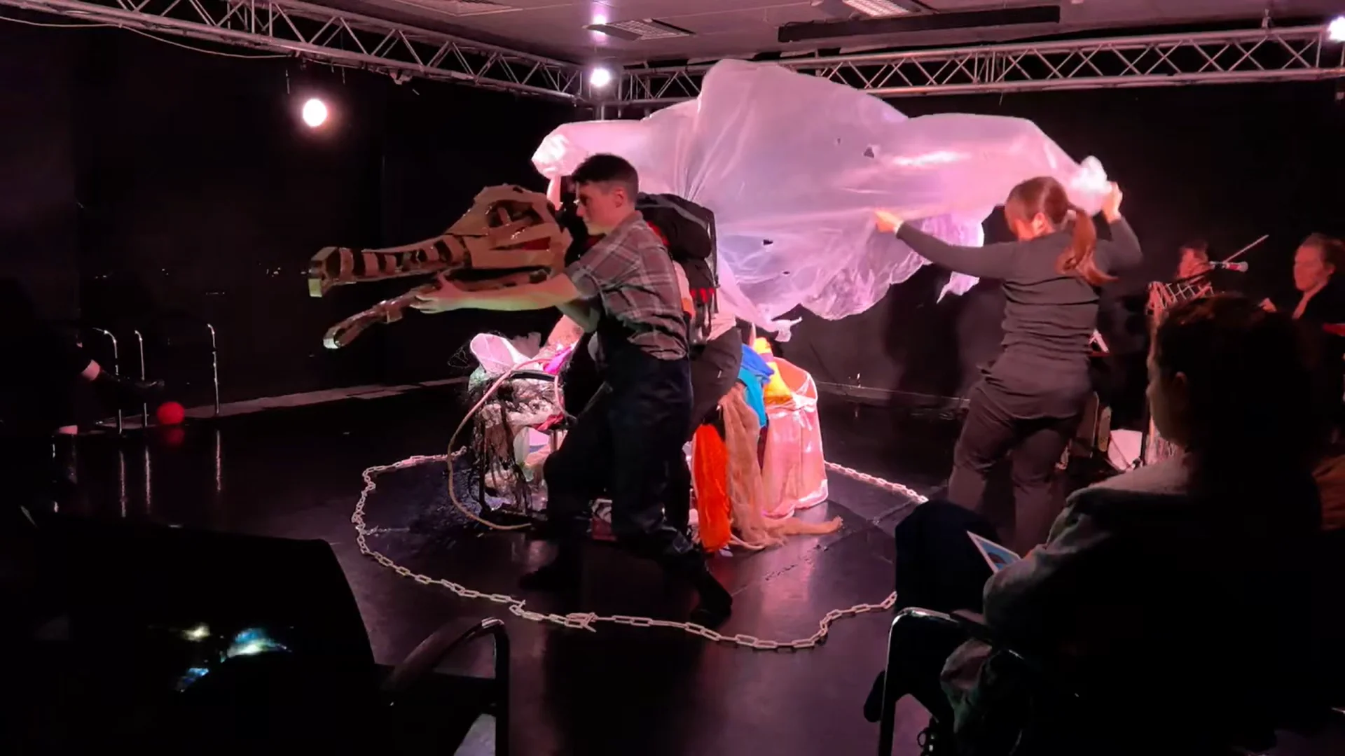 A performer holds a large cardboard dinosaur head while another waves a large plastic sheet on a stage surrounded by a chain.